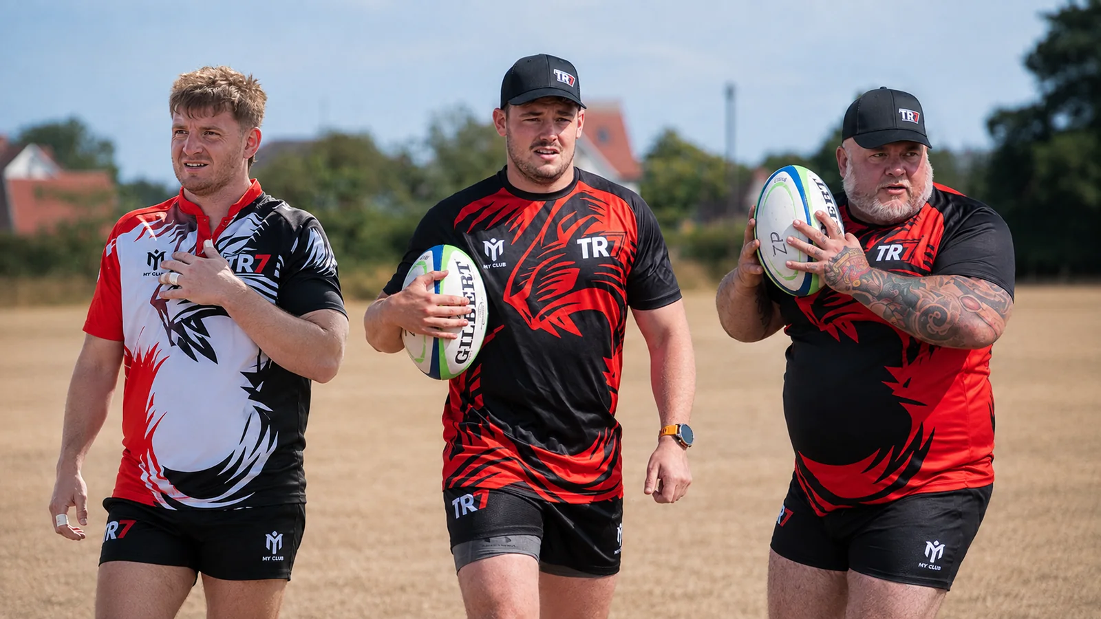 Three TR7 players walking across a sun-bleached pitch carrying rugby balls, two in TR7 caps
